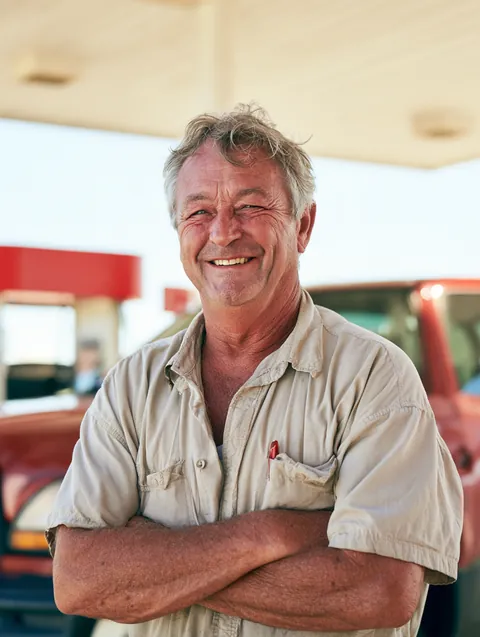Grinning older man standing under a bright gas station canopy with his pickup truck parked behind him.