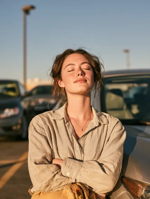 Young woman exhaling with relief beside her compact car in a sunny parking lot after sorting out a $500 loan.