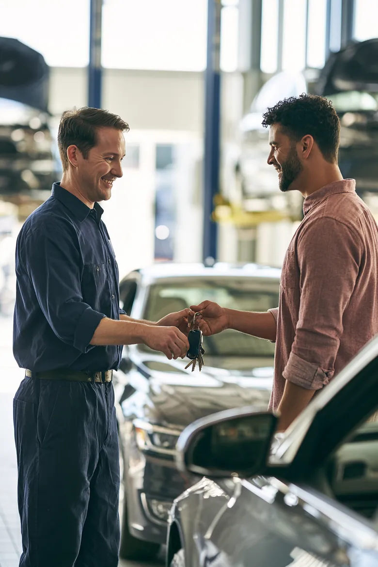 A mechanic in a navy work shirt hands car keys back to a man beside his freshly serviced sedan in a bright repair shop.