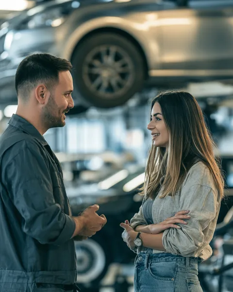 A mechanic explains a repair to a young woman while her car sits on a lift in a bright shop.