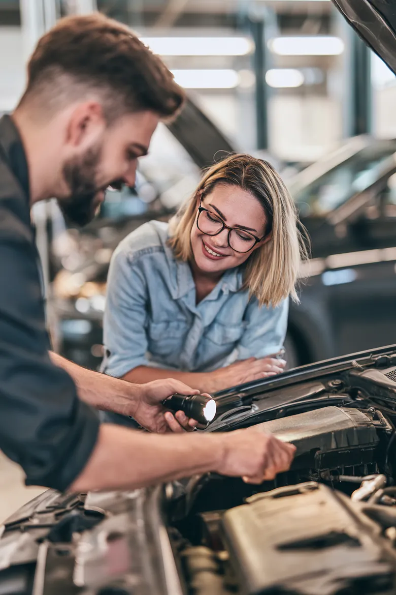 A mechanic points a flashlight at an engine bay while explaining a repair to a woman weighing an Auto Approve loan request.