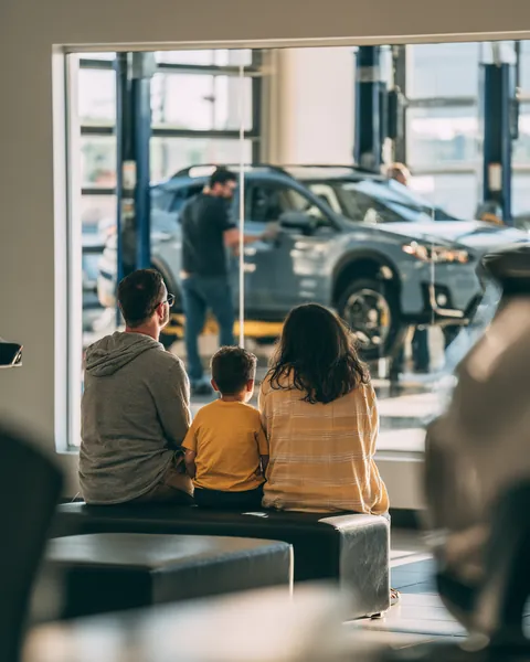 A family with a young son watches through a tire shop's glass wall as technicians mount new wheels on their crossover.