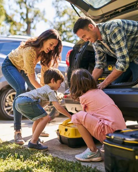 Family in their 30s packing a roadside emergency kit into their SUV trunk with their kids helping