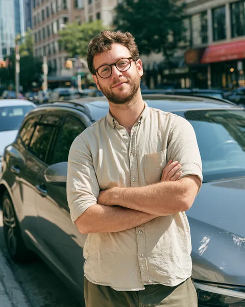 Man in his 30s shrugging beside his car with a small bumper scratch on a city street