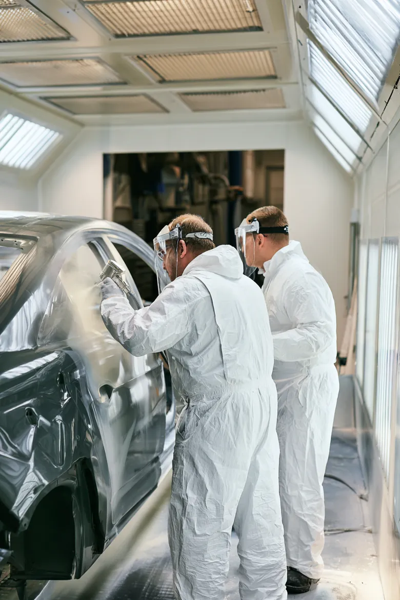 Painter spraying a car panel in a paint booth while a colleague checks the finish, from the Auto Approve fender bender guide