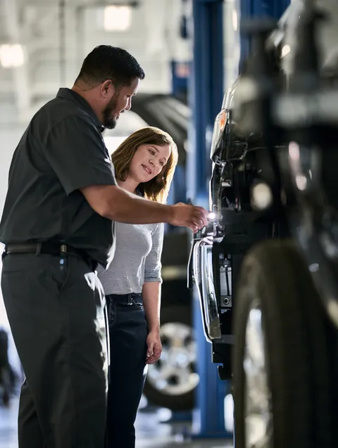 A technician tests headlight alignment at a modern inspection station while a woman in her thirties looks on.